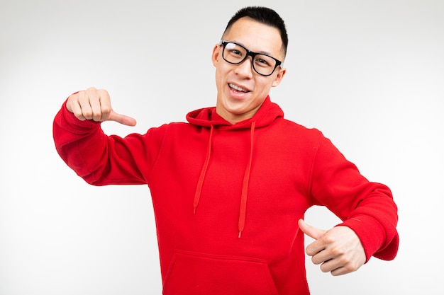 Schöne brünette asiatische in rotem Pullover und Brille für Sehvermögen zeigt Finger auf einem Pullover auf einem weißen Hintergrund