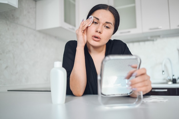 Schöne brünette Frau, die Make-up von ihrem Gesicht mit Spiegel entfernt. Nettes schönes natürliches Schönheitsmädchen, das Gesicht mit Baumwollschwamm reinigt. Kosmetologie und Spa, Problemhautpflege, Aknebehandlungskonzept