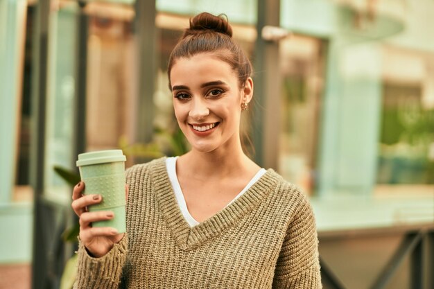 Schöne brünette Frau lächelt glücklich und selbstbewusst im Freien in der Stadt an einem sonnigen Herbsttag und trinkt eine Tasse Kaffee zum Mitnehmen