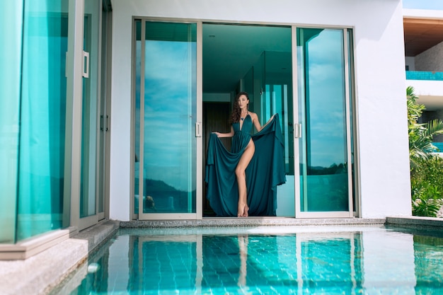 Schöne Brünette im blauen Abendkleid, das nahe am Pool in einer Villa stehend aufwirft