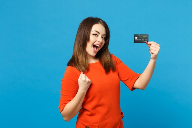 Schöne brünette junge Frau in rot-orangefarbenem Kleid hält in der Hand Kreditkarte isoliert über trendigem blauem Wandhintergrund, Studioportrait. Lifestyle-Modekonzept für Menschen. Mock-up-Kopienbereich.