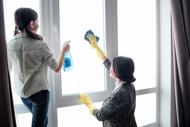 Schöne brünette kaukasische Mutter und Tochter, die Fenster zusammen im Raum reinigen. Mädchen benutzen Spray. Junge Frau, die Fenster wäscht. Sie arbeiten zusammen.