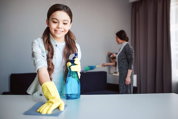 Schöne brünette kaukasische Mutter und Tochter, die zusammen im Raum reinigen. Mädchen halten Spray in Händen und sauberen Tisch. Sie trägt gelbe Handschuhe. Ihre Mutter sprengt den Staub dahinter.