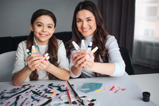 Schöne brünette kaukasische Mutter und Tochter malen zusammen im Raum. Fröhliche glückliche Menschen schauen vor die Kamera und lächeln. Sie halten weiße Röhren mit Farbe in den Händen. Pinsel und Palette auf dem Tisch.
