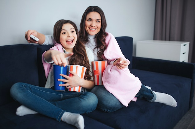 Schöne brünette kaukasische Mutter und Tochter sitzen zusammen im Raum. Emotional aufgeregtes Mädchen zeigen nach vorne. Sie hält Korbpopcorn und Cola. Mutter lächelt und benutzt die Fernbedienung.