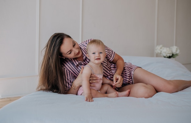 Schöne brünette Mama im Pyjama liegt mit einem blonden Baby im Schlafzimmer auf dem Bett