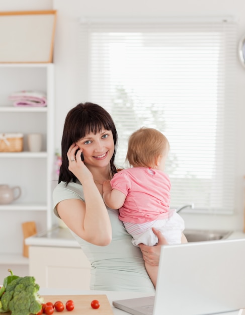 Schöne Brunettefrau am Telefon beim Halten ihres Babys in ihren Armen