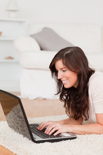 Schöne Brunettefrau, die mit ihrem Laptop beim Lügen auf einem Teppich sich entspannt