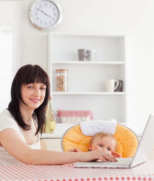 Schöne Brunettefrau, die mit ihrem Laptop nahe bei ihrem Baby beim Sitzen sich entspannt