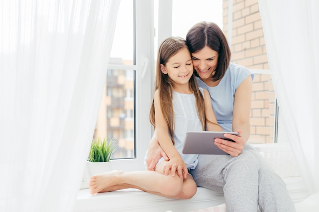 Schöne Brunettemutter und -tochter verbringen Freizeit zusammen, sitzen auf Fensterbrett, passen interessanten Film über digitale Tablette auf