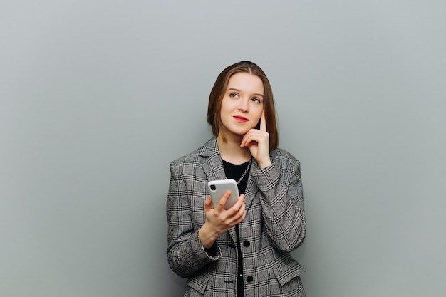 Schöne Büroangestellte in einer Jacke steht auf dem Hintergrund einer grauen Wand mit einem Smartphone