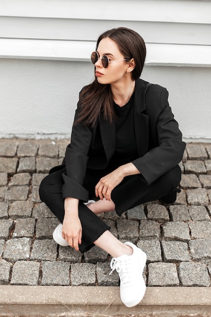 Schöne coole junge Frau in trendiger Sonnenbrille in cooler schwarzer Jugendkleidung in weißen trendigen Turnschuhen entspannt sich auf Steinfliesen in der Nähe des Vintage-Gebäudes in der Stadt. Modisches Mädchen im stilvollen Outfit genießt Ruhe.