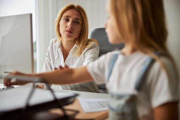 Schöne Dame, die am Computer arbeitet, wo sie dem weiblichen Kind verbietet, Dokumente auf dem Schreibtisch zu berühren