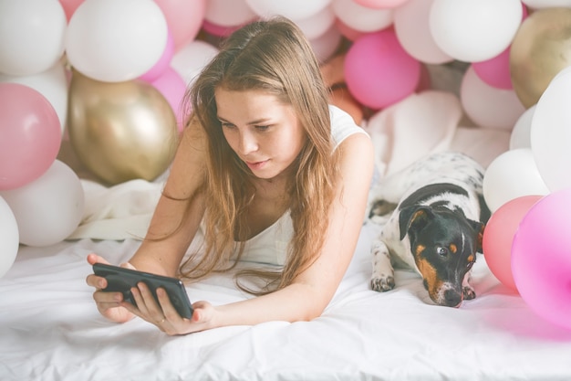 Schöne Dame im Pyjama, die Selfie in ihrem Schlafzimmer mit Telefon macht und ihren Hund umarmt.