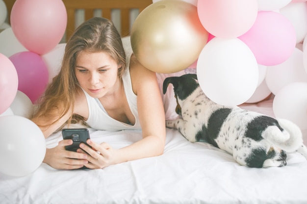 Schöne Dame im Pyjama, die Selfie in ihrem Schlafzimmer mit Telefon macht und mit ihrem Hund rumhängt.