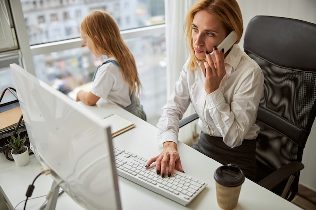 Schöne Dame in Geschäftskleidung, die am Computer arbeitet und im Büro mit dem Handy telefoniert