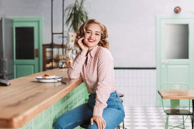 Schöne Dame in Hemd und Jeans, die mit einem Stück Kuchen in der Nähe an der Theke sitzt und freudig in die Kamera blickt, während sie Zeit im Café verbringt