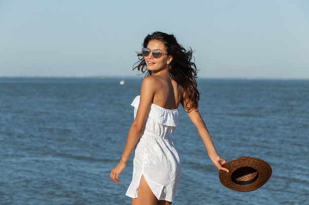 Schöne dunkelhaarige Mädchen in weißem Kleid und Sonnenbrille halten Hut in der Hand und gehen an einem sonnigen Tag in der Nähe des Meeres. .