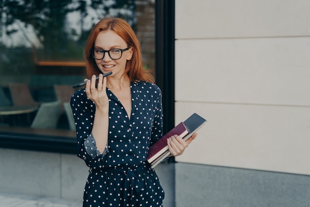 Schöne elegante lächelnde Geschäftsfrau im Kleid mit Freisprecheinrichtung oder virtuellem Sprachassistent auf dem Handy, während sie mit Laptop in der Hand im Freien steht, zeichnet Audionachricht für Kollegen auf