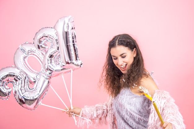 Schöne emotionale junge Brünette mit lockigem Haar festlich gekleidet, die eine Feuerwerkskerze in ihrer Hand und silberne Luftballons für das Neujahrskonzept auf einer rosa Wand hält