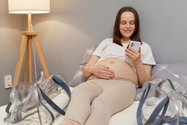 Schöne, entspannte schwangere Frau liegt auf dem Bett, berührt ihren nackten Bauch mit dem Smartphone, scrollt online, surft auf Internetseiten und liest Informationen über die Geburt