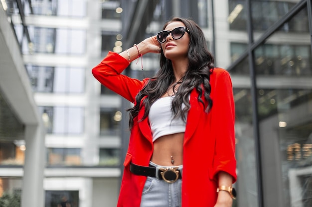 Schöne, erfolgreiche, modische Geschäftsfrau mit lockigem Haar und modischer Sonnenbrille, die eine modische rote Blazer-Top-Jeans und eine Handtasche trägt, geht in der Stadt in der Nähe moderner Gebäude spazieren