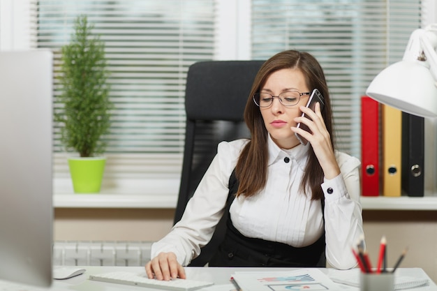 Schöne ernsthafte Geschäftsfrau in Anzug und Brille, die am Schreibtisch sitzt, an einem modernen Computer mit Dokumenten im hellen Büro arbeitet, auf dem Handy spricht, Probleme löst, beiseiteschaut