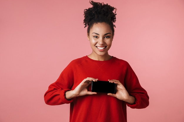 schöne erstaunliche junge afrikanische frau, die isoliert über rosa wand posiert und die anzeige des handys zeigt.