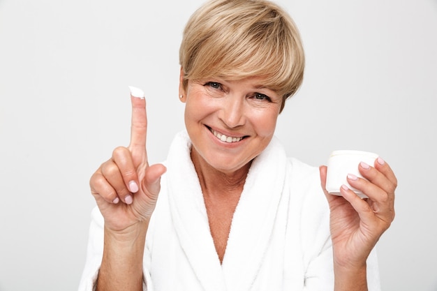 Schöne erwachsene Frau mit kurzen blonden Haaren, die einen weißen Hausmantel trägt, der ein Gesichtscremeglas isoliert über der weißen Wand hält