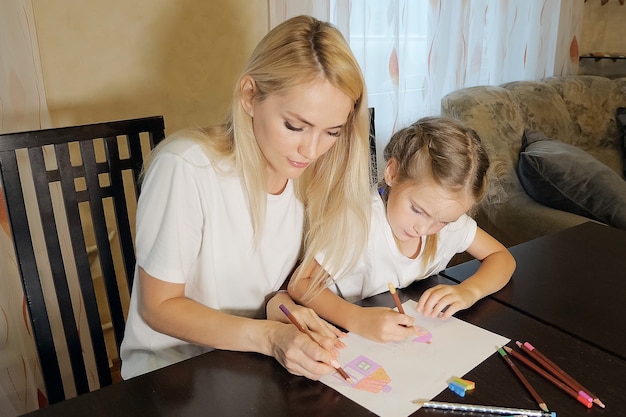 Schöne erwachsene Frau und Mädchen malen mit Bleistiften auf Papier am Tisch zu Hause sitzen