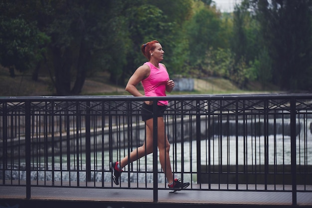 Schöne erwachsene rothaarige Sportlerin in rosa Top und Shorts macht einen morgendlichen Lauf auf der Stadtbrücke über den Fluss. Gesunder Lebensstil, Joggen