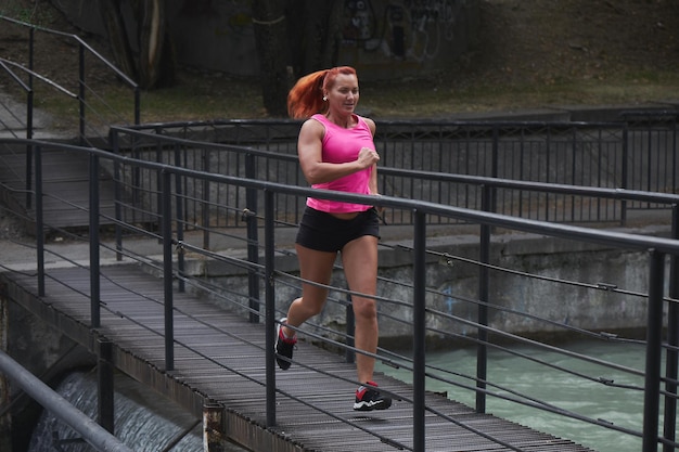 Schöne erwachsene rothaarige Sportlerin in rosa Top und Shorts macht einen morgendlichen Lauf auf der Stadtbrücke über den Fluss. Gesunder Lebensstil, Joggen