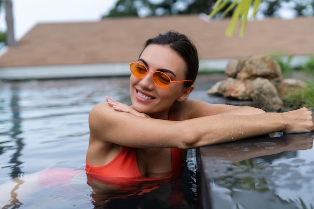 Schöne europäische Frauen in zweiteiligem orangefarbenem Bikini-Badeanzug, gebräunt mit trendiger Sonnenbrille draußen am tropischen Swimmingpool