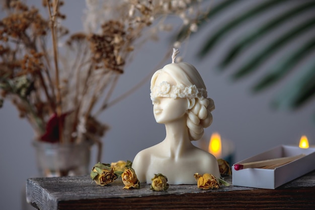 Schöne feminine handgefertigte Kerze in Form eines weiblichen Körpers auf einem Holzständer. In der Nähe befinden sich Streichhölzer und gelbe Rosenknospen