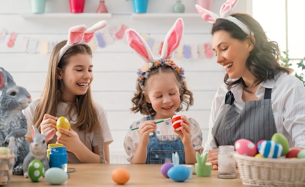 Schöne Ferien! Eine Mutter und ihre Töchter bemalen Eier. Familie bereitet sich auf Ostern vor. Nette kleine Kindermädchen tragen Hasenohren.