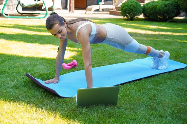 Schöne Fitness-Frau, die eine Plankenübung macht und sich Online-Tutorials zum Laptop-Training im Freien ansieht. Gesunder Lebensstil. Mädchen macht Sport im Garten