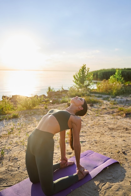 Schöne, fitte Dame in Sportleggings und bauchfreiem Top, die an einem sonnigen Tag am Strand Yoga praktiziert