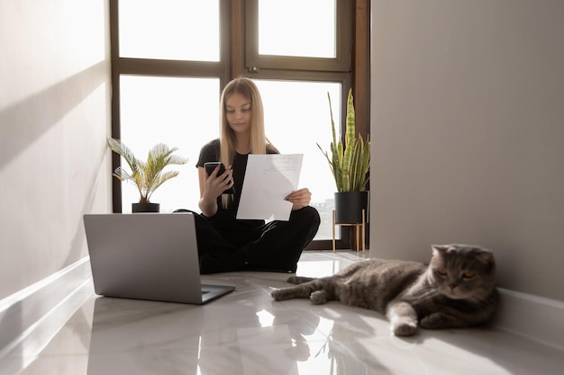 Schöne Frau arbeitet zu Hause am Computer, der am Fenster auf dem Boden sitzt, und prüft Dokumente und Benachrichtigungen am Telefon