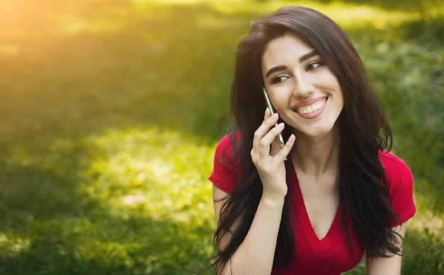 Schöne Frau, die auf Gras im Park sitzt und auf Smartphone spricht, Kopienraum