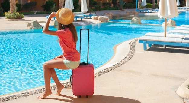 Schöne Frau, die auf rosa Koffer nahe Poolpoolbereich des Schwimmhotels sitzt.