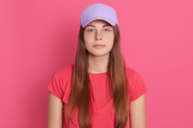 Schöne Frau, die Baseballmütze und T-Shirt trägt, die lokal über rosa Wand mit ernstem Ausdruck auf Gesicht aufwirft, das langes glattes Haar hat.