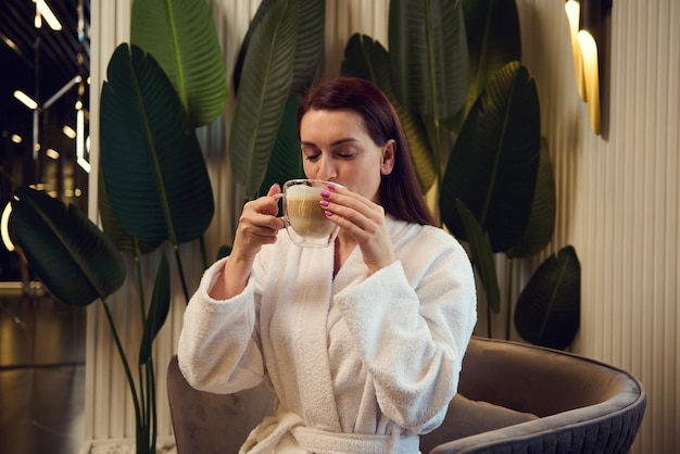 Schöne Frau, die Cappuccino trinkt, sitzt auf einem Sessel in einem Lounge-Bereich eines Spa-Salons Attraktive entspannte europäische hübsche Frau, die eine Kaffeepause genießt, während sie eine luxuriöse Wellness-Spa-Klinik besucht