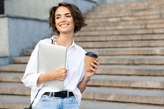 Schöne Frau, die draußen geht und Laptop und Kaffeetasse hält