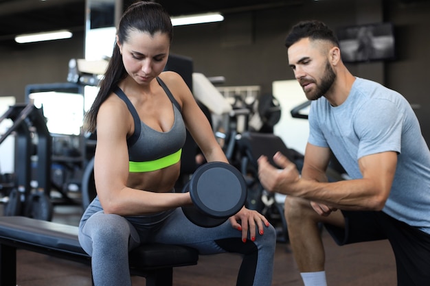 Schöne Frau, die eine Übung mit Gewicht unter Anleitung eines Trainers im Fitnessstudio macht.