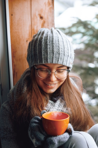 Schöne frau, die eine tasse kaffee oder kakao in handschuhen hält und trinkt, die nach hause am fenster sitzen. unscharfer winterschneebaumhintergrund.