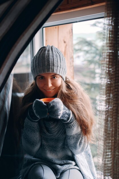 Schöne Frau, die eine Tasse Kaffee oder Kakao in Handschuhen hält und trinkt, die zu Hause am Fenster sitzen