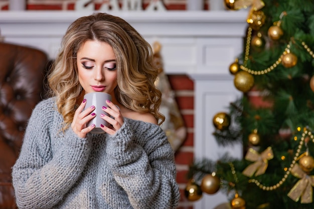 Schöne Frau, die eine Tasse warmen Kaffee in ihren Händen auf dem Hintergrund eines weißen Kamins im Raum mit Neujahrsdekorationen und einem Weihnachtsbaum hält