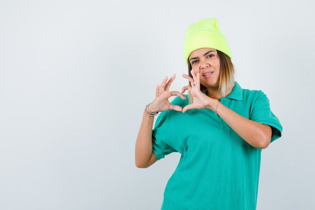 Schöne Frau, die Herzgeste in Polo-T-Shirt, Mütze zeigt und selbstbewusst aussieht. Vorderansicht.