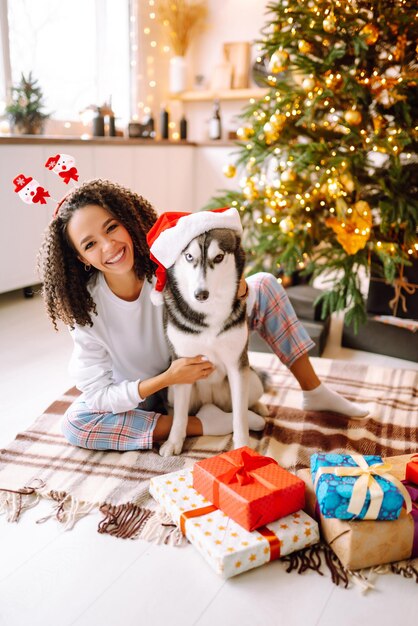 Schöne Frau, die mit ihrem Hund spielt und Spaß hat, während sie neben dem Weihnachtsbaum sitzt