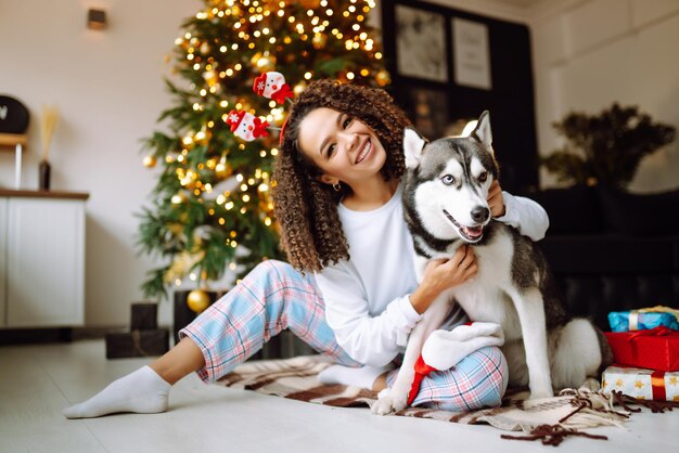 Schöne Frau, die mit ihrem Hund spielt und Spaß hat, während sie neben dem Weihnachtsbaum sitzt